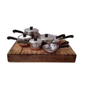 May include: A collection of stainless steel cookware featuring copper-toned bases and black handles. The set includes various pots and pans with matching lids, arranged together to display the metallic finish and the dark, heat-resistant handles.
