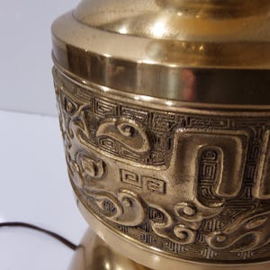 May include: Close-up of a brass lamp base with intricate embossed designs. The lamp base has a detailed pattern of swirling shapes and geometric elements. The lamp is a warm gold color and has a classic, ornate style.