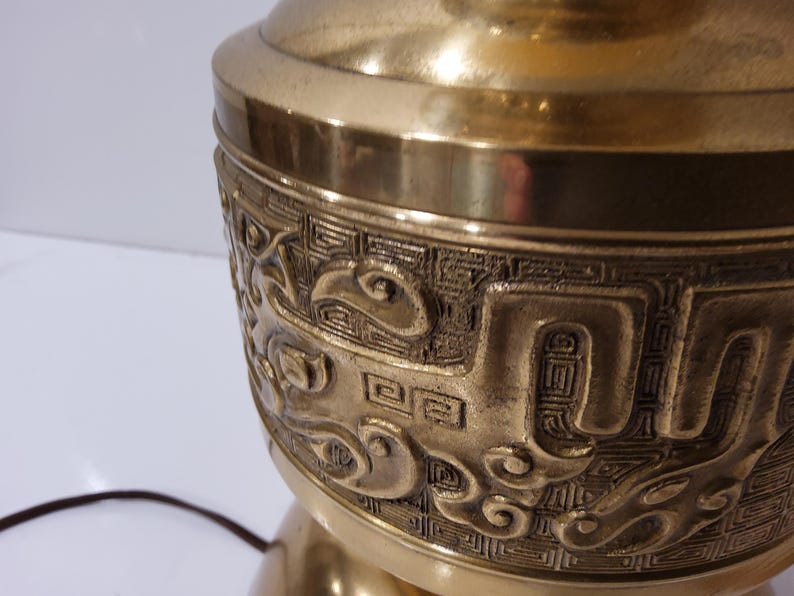 May include: Close-up of a brass lamp base with intricate embossed designs. The lamp base has a detailed pattern of swirling shapes and geometric elements. The lamp is a warm gold color and has a classic, ornate style.