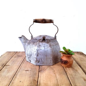 May include: A vintage metal teapot with a wooden handle and a small decorative pot with a green plant. The teapot has a unique, geometric shape and sits on a rustic wooden surface.