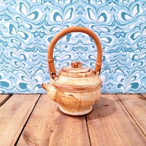 May include: A ceramic teapot with a light brown glaze and a wooden handle. The teapot has a rounded body, a spout, and a lid. The handle is made of a curved piece of wood. The background is a blue and white patterned design.