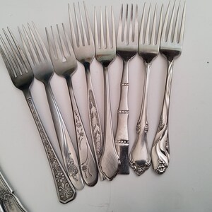 May include: A collection of eight silver forks with different decorative patterns on the handles. The forks are arranged in a semi-circle on a white surface.