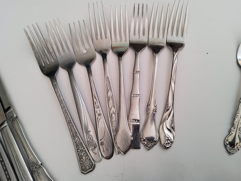 May include: A collection of eight silver forks with different decorative patterns on the handles. The forks are arranged in a semi-circle on a white surface.