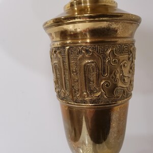 May include: A brass-colored decorative vase with an ornate, embossed design around the middle. The vase has a flared top and a rounded base. The embossed design features intricate patterns and shapes. The vase is made of metal and has a shiny finish.