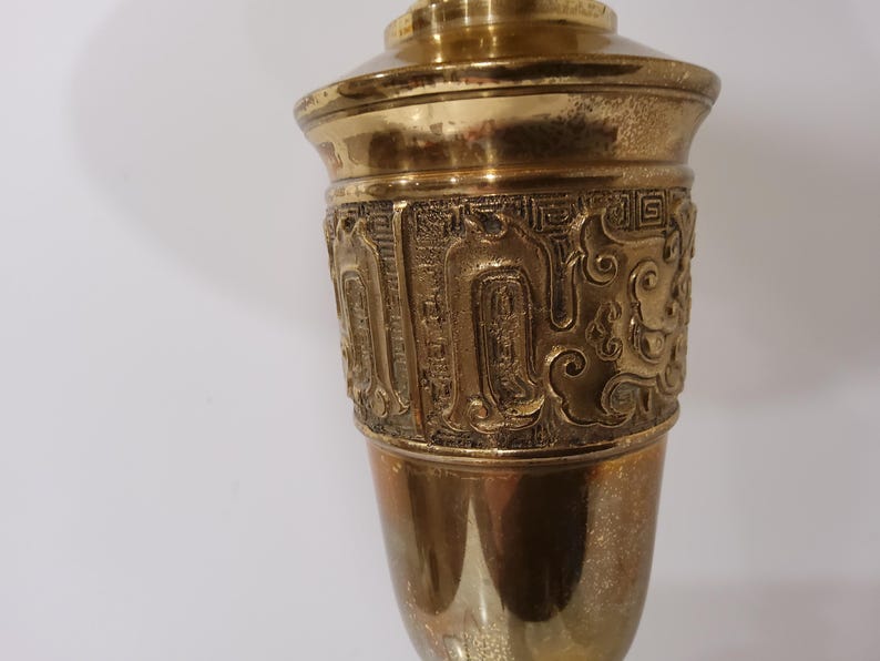 May include: A brass-colored decorative vase with an ornate, embossed design around the middle. The vase has a flared top and a rounded base. The embossed design features intricate patterns and shapes. The vase is made of metal and has a shiny finish.