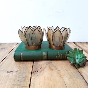 May include: Two lotus-shaped candle holders constructed from translucent, off-white shell petals with copper-toned metal bases. The holders are positioned side-by-side, each featuring a layered, petal-like design that mimics the appearance of a flower.