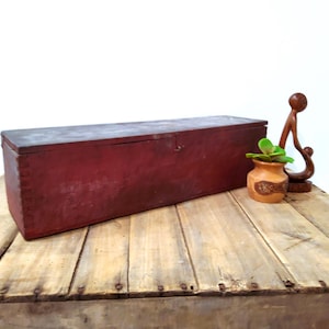 May include: A long, rectangular, red wooden box with a hinged lid sits on a rustic wooden surface. A small terracotta pot with a succulent and a wooden sculpture are placed nearby. The box appears aged.