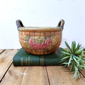 May include: A round ceramic basket-weave planter featuring two side handles and a central relief design of pink roses with green leaves. The exterior has a warm tan and brown glazed finish with a textured pattern resembling woven wicker material.