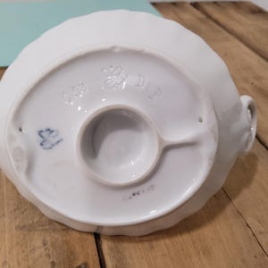 May include: The underside of a white ceramic object featuring a scalloped edge and a central circular base. Embossed markings include a crown symbol, the letters C.S. and DeP., and a small blue painted mark. The surface is smooth and glossy.