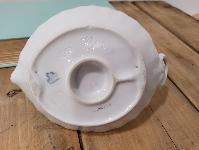 May include: The underside of a white ceramic object featuring a scalloped edge and a central circular base. Embossed markings include a crown symbol, the letters C.S. and DeP., and a small blue painted mark. The surface is smooth and glossy.