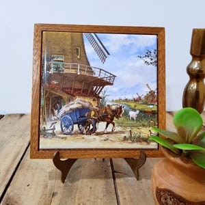 May include: A framed ceramic tile featuring a pastoral scene with a windmill, horse-drawn cart, and figures. The artwork is set against a blue sky with other windmills in the distance. The frame is made of wood.