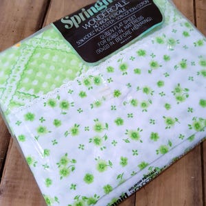 Puede incluir: Una sábana plana tamaño queen empaquetada con un estampado floral. La sábana es blanca con pequeñas flores verdes y un borde con estampado verde. El embalaje incluye la marca "Springmaid" y detalles del producto.