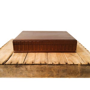 May include: A dark brown wooden box with a carved design, placed on a light-coloured, weathered wooden platform. The box has a rectangular shape and a flat top. The platform appears to be made of reclaimed wood.