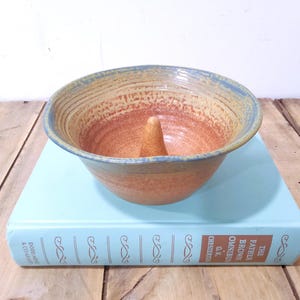 May include: A handmade ceramic apple baker bowl with a central core. The bowl features a textured surface with a gradient of colors, including orange, tan, and blue. It sits on a light blue book with gold lettering.