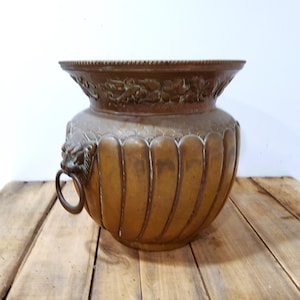 May include: A bronze-colored, antique-style planter with a wide, decorative rim and a bulbous body. The planter features vertical ribbed detailing and lion head handles with rings. The rim has a floral design.