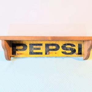 May include: Wooden shelf with a yellow and black Pepsi sign. The shelf has two hooks on the front.