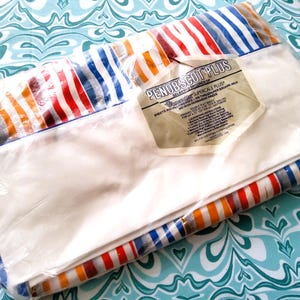 K&ouml;nnte beinhalten: Ein verpacktes Set wei&szlig;er Bettlaken mit einem gestreiften Rand in Rot, Orange, Blau und Grau. Die Verpackung enth&auml;lt ein Etikett mit der Aufschrift "PENOBSCOT PLUS". Die Laken liegen auf einer t&uuml;rkisfarbenen und wei&szlig;en Oberfl&auml;che.