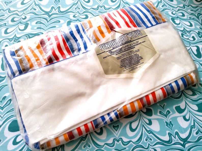 K&ouml;nnte beinhalten: Ein verpacktes Set wei&szlig;er Bettlaken mit einem gestreiften Rand in Rot, Orange, Blau und Grau. Die Verpackung enth&auml;lt ein Etikett mit der Aufschrift "PENOBSCOT PLUS". Die Laken liegen auf einer t&uuml;rkisfarbenen und wei&szlig;en Oberfl&auml;che.