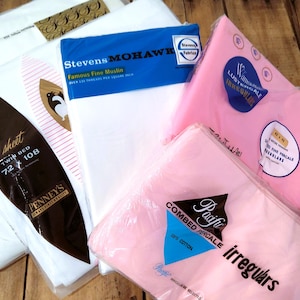 May include: An assortment of vintage bed sheets in various colors, including white and pink. Several packages are visible, with labels indicating the brand and type of fabric, such as "Famous Fine Muslin" and "Combed Percale".
