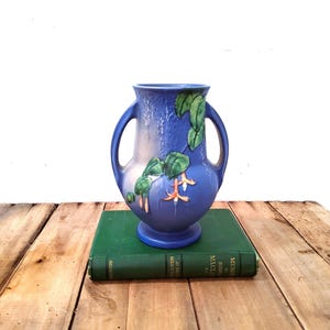 May include: A blue ceramic vase with two handles and a pedestal base, featuring a raised relief design of green leaves and orange fuchsia flowers. The vase has a smooth, glossy finish with a gradient of light and dark blue tones across its surface.