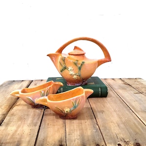 May include: A ceramic teapot with a high arched handle and a matching lid, accompanied by two small matching open bowls. The set features a warm orange glaze with embossed green leaf and white floral details on the exterior of each piece.
