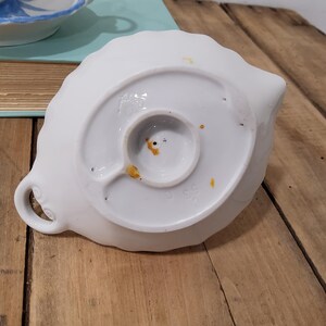 May include: A white ceramic dish shaped like a leaf with a scalloped edge and a small circular handle. The underside features a central circular base, a small loop, and visible orange-brown discoloration spots on the white glazed surface.