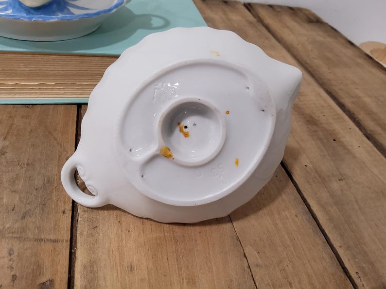 May include: A white ceramic dish shaped like a leaf with a scalloped edge and a small circular handle. The underside features a central circular base, a small loop, and visible orange-brown discoloration spots on the white glazed surface.