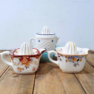 May include: Three ceramic citrus juicers with white reamer tops. One juicer features a white lattice pattern with blue accents. The other two juicers have square bases with floral patterns in orange, red, blue, and yellow colors, each with a handle.