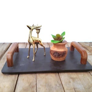 May include: A decorative tray with wooden handles, holding a gold deer figurine with a candle holder, and a small terracotta pot with a faux succulent. The tray is dark gray, and the handles are a warm brown.
