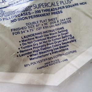 K&ouml;nnte beinhalten: Nahaufnahme eines wei&szlig;en Lakens in der Originalverpackung. Die Verpackung enth&auml;lt Text, der besagt, dass es sich um ein doppeltes Flachlaken mit den Ma&szlig;en 206 x 244 cm handelt und aus 50 % Polyester und 50 % Baumwolle besteht.