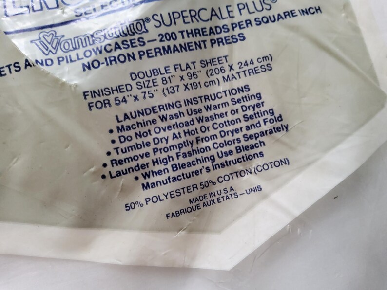 K&ouml;nnte beinhalten: Nahaufnahme eines wei&szlig;en Lakens in der Originalverpackung. Die Verpackung enth&auml;lt Text, der besagt, dass es sich um ein doppeltes Flachlaken mit den Ma&szlig;en 206 x 244 cm handelt und aus 50 % Polyester und 50 % Baumwolle besteht.