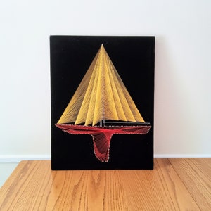 May include: A black framed wall art piece with a yellow and red string art design of a sailboat. The sailboat is made of string stretched between nails on the black background.