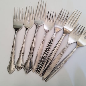 May include: A collection of eight silver forks with various ornate designs on the handles. The forks are arranged in a fan shape on a white surface.