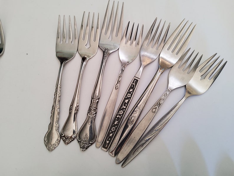 May include: A collection of eight silver forks with various ornate designs on the handles. The forks are arranged in a fan shape on a white surface.