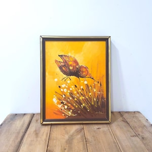 May include: Framed vintage painting featuring a butterfly with brown and orange wings, hovering over a field of white and yellow flowers. The artwork is set against a warm yellow background and framed in a gold-colored frame.