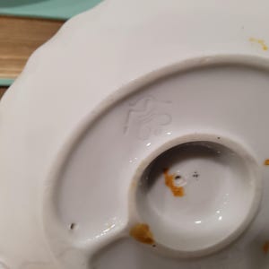 May include: The underside of a white ceramic object featuring a scalloped edge and a circular base. A stamped maker's mark is visible on the surface, along with small orange-brown stains and two tiny holes near the center of the circular base.