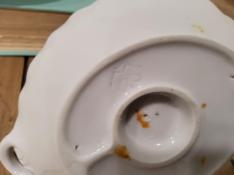May include: The underside of a white ceramic object featuring a scalloped edge and a circular base. A stamped maker's mark is visible on the surface, along with small orange-brown stains and two tiny holes near the center of the circular base.