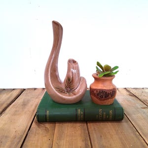 May include: A light brown ceramic vase with a unique curved design and a small, round opening. Next to it is a small, orange-brown pot with a plant and a green book. The objects are on a wooden surface.