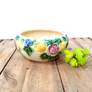 May include: A round ceramic planter with a cream-colored base featuring a raised, embossed floral pattern. The design includes painted yellow, pink, and blue flowers with green leaves encircling the exterior. A small cluster of bright green faux flowers sits beside the planter.