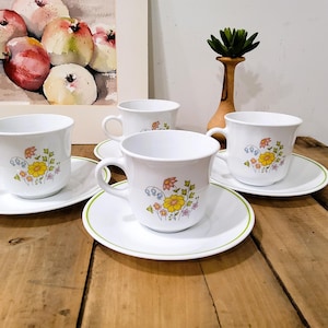 May include: A set of four white teacups and saucers with a floral design. Each cup and saucer has a green trim. The cups and saucers are arranged on a wooden surface. A watercolor painting of apples is in the background.