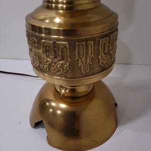 May include: A vintage brass table lamp with a decorative design. The lamp features a bulbous base and a detailed band of embossed patterns. The lamp has a warm, golden hue and a classic aesthetic, suitable for home decor.