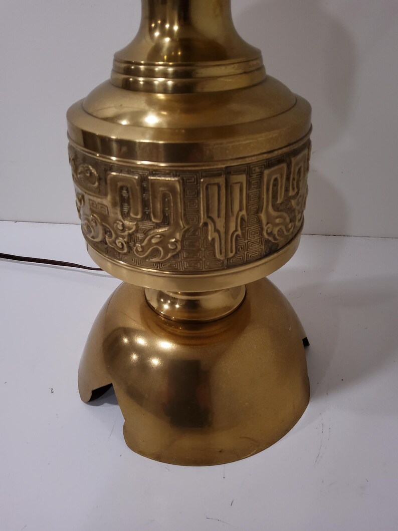 May include: A vintage brass table lamp with a decorative design. The lamp features a bulbous base and a detailed band of embossed patterns. The lamp has a warm, golden hue and a classic aesthetic, suitable for home decor.
