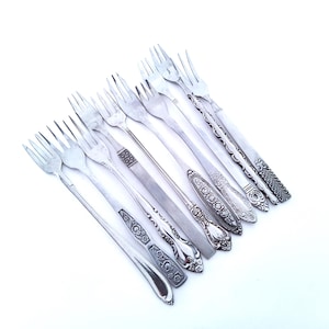 May include: An assortment of silver forks with various handle designs, arranged diagonally against a white background. The forks showcase different patterns, from simple to ornate, reflecting a vintage or antique aesthetic.