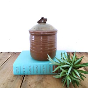 May include: A cylindrical brown ceramic jar with a matching lid featuring a small sculpted handle on top. The jar has a smooth, glossy finish with subtle horizontal ridges along the body, showcasing a warm, earthy tone and a slightly lighter lid.