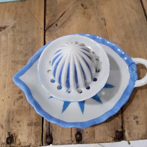 May include: A white ceramic citrus juicer featuring a central reamer with blue vertical stripes, surrounded by a circular strainer plate with drainage holes, all resting on a leaf-shaped base with a blue painted edge and a small handle.