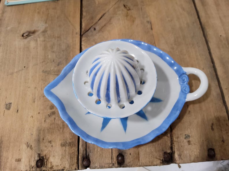 May include: A white ceramic citrus juicer featuring a central reamer with blue vertical stripes, surrounded by a circular strainer plate with drainage holes, all resting on a leaf-shaped base with a blue painted edge and a small handle.