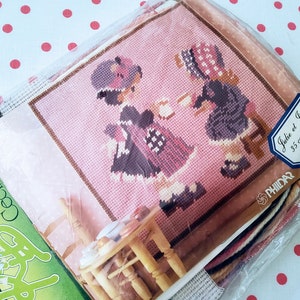 May include: A pink needlepoint kit featuring two girls in dresses, one sitting on a chair and the other standing. The kit includes a printed canvas, yarn, and instructions. The kit is labeled "Julie et Juliette 35 cm x 35 cm 903.11".