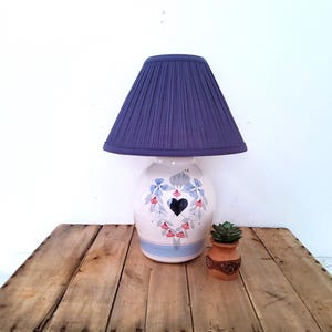 May include: A table lamp with a dark purple pleated fabric shade and a white ceramic base. The base features a painted design of blue flowers, green leaves, and a central black heart, with a light blue horizontal band near the bottom.