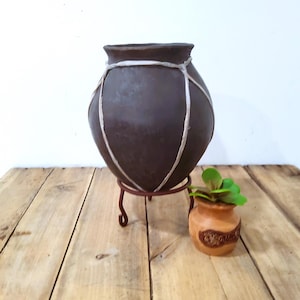 May include: A dark brown, round ceramic pot with a white rope design, sitting on a small, dark brown metal stand. A small, light brown pot with a green plant is next to it. The objects are on a wooden surface.
