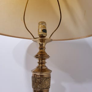 May include: A vintage brass table lamp with a beige lampshade. The lamp features an ornate base with a detailed floral pattern. The lampshade is a soft, neutral color, and the lamp is designed for indoor use.